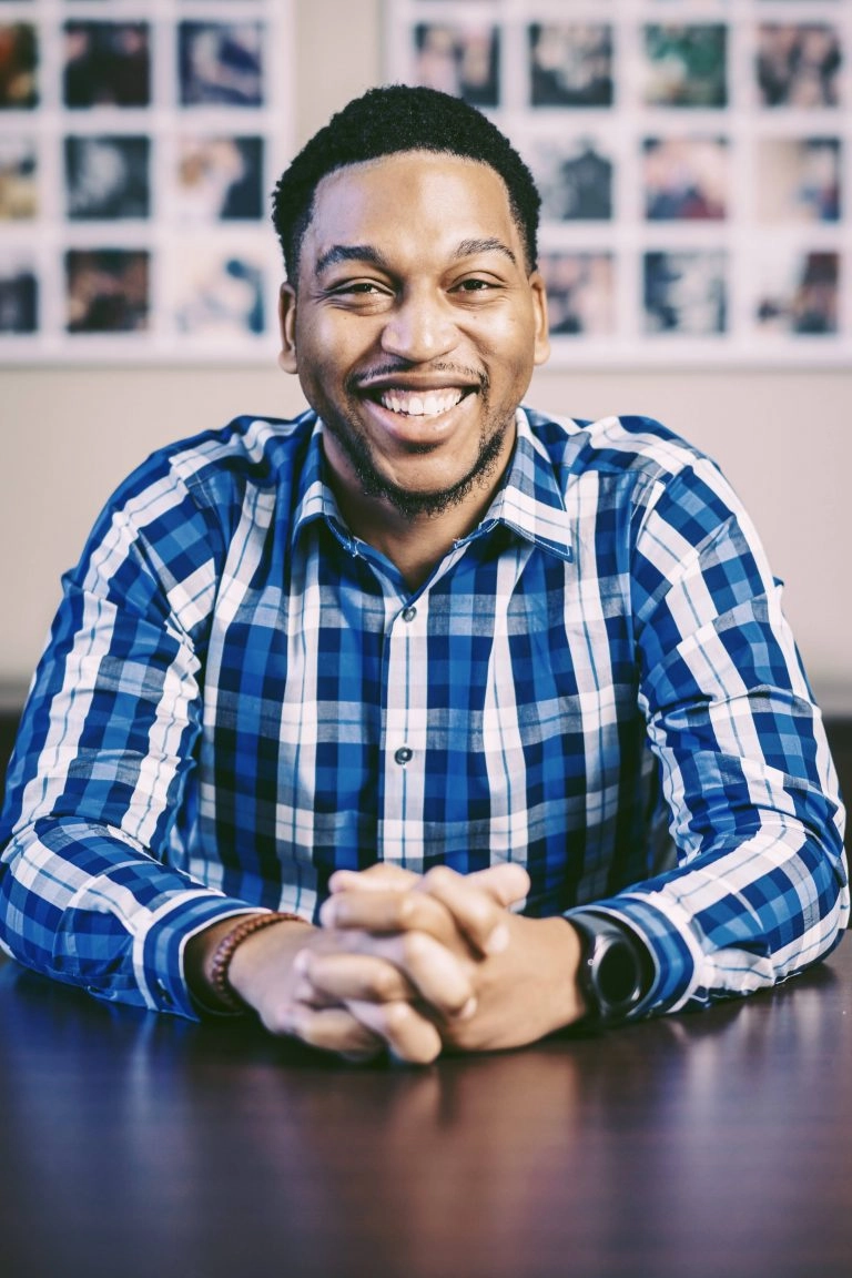 Smiling man in a blue plaid shirt sitting at a table with hands clasped, against a blurred background of photos. Howard Creative Co
