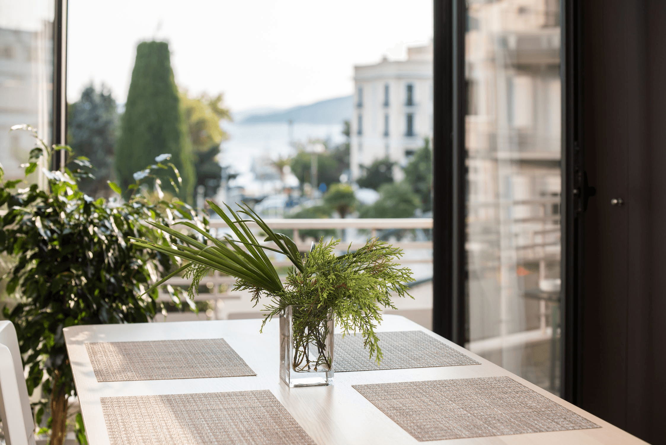 A sunlit dining table with a vase of greenery and four placemats is near a window overlooking a cityscape and distant water. Howard Creative Co