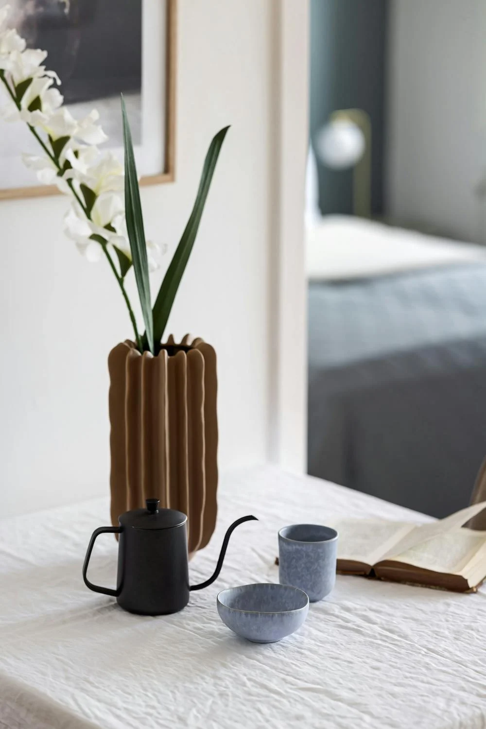 A table with a ceramic vase holding white flowers, a black teapot, a blue cup, a bowl, and an open book, set against a blurred background of a bed and lamp. Howard Creative Co